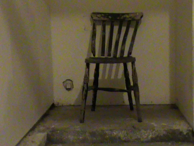A crooked wooden chair sits in a small, grimy room.